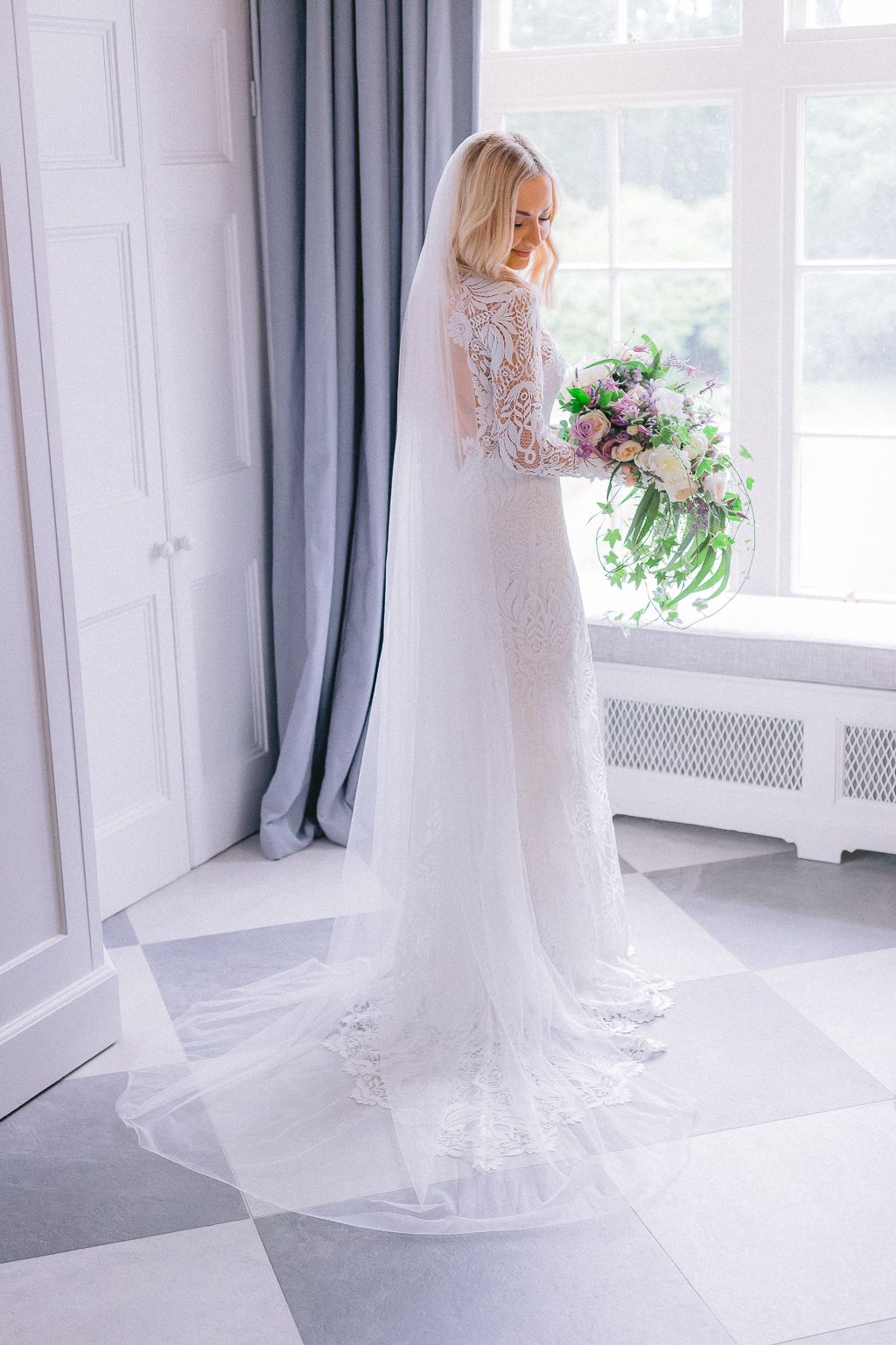 Bride stands ready for wedding facing window holding bouquet of flowers